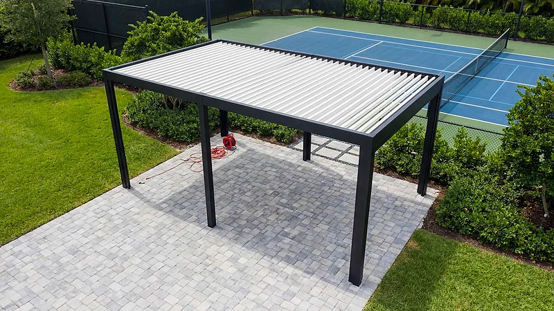 Black-framed louvered roof with closed white blades over a paver patio, seen from above beside a hedge and a tennis court