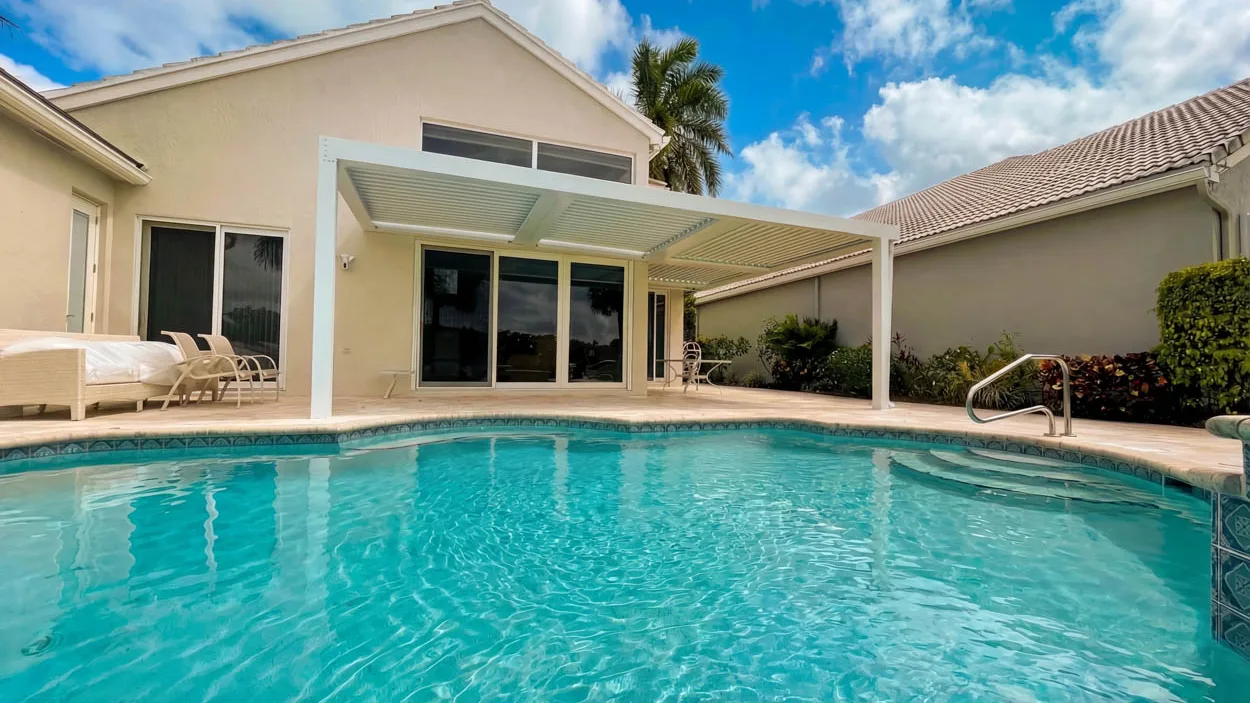 White louvered roof over a covered patio on the far side of a pool, with lounge chairs on the deck to the left and a handrail at the pool steps on the right.