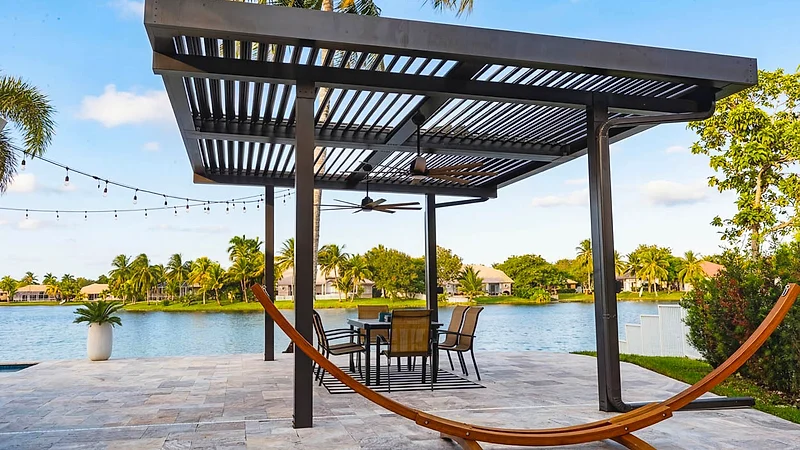 Dark-framed louvered roof with a ceiling fan over a dining set on a stone patio by a lake, with string lights and a hammock