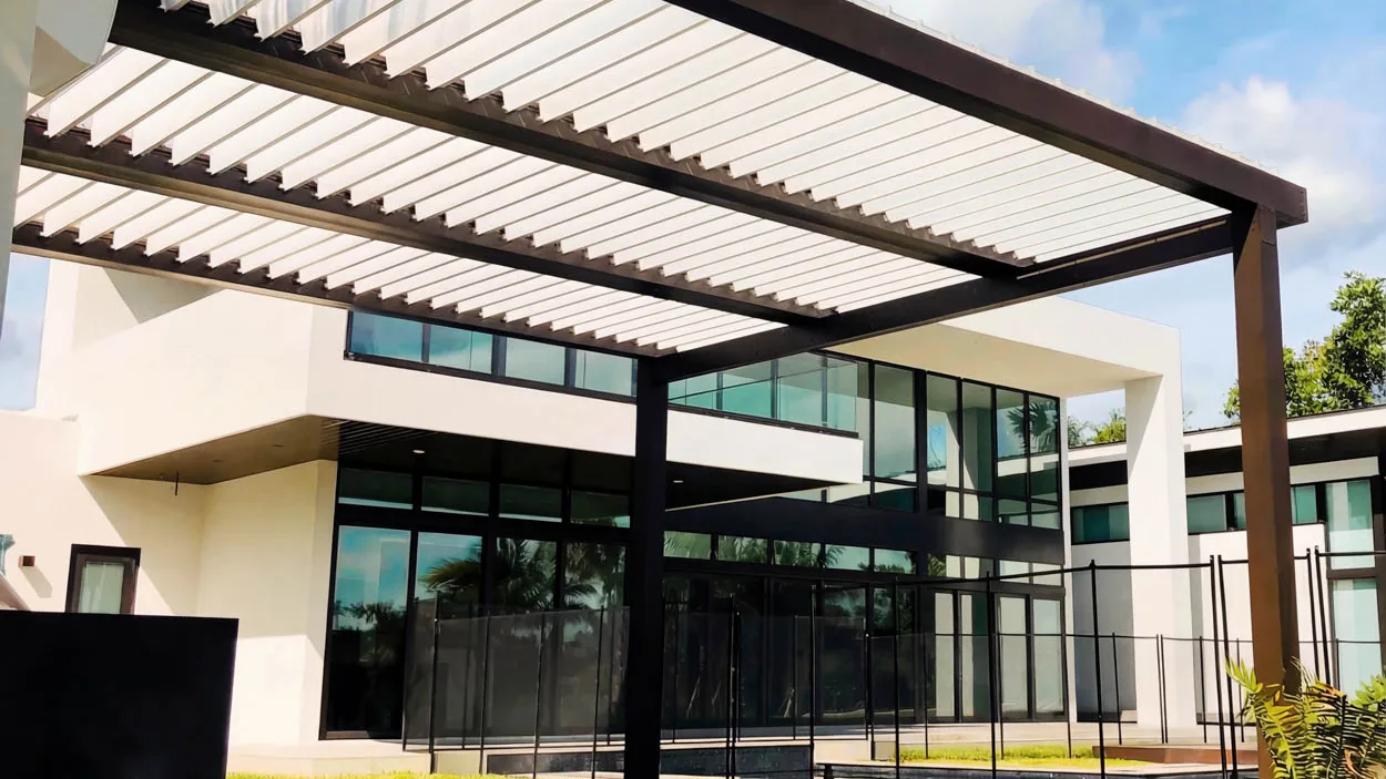 Louvered roof with white blades tilted partly open in a dark bronze frame, attached to a modern glass-walled house over a concrete patio behind a black pool fence.