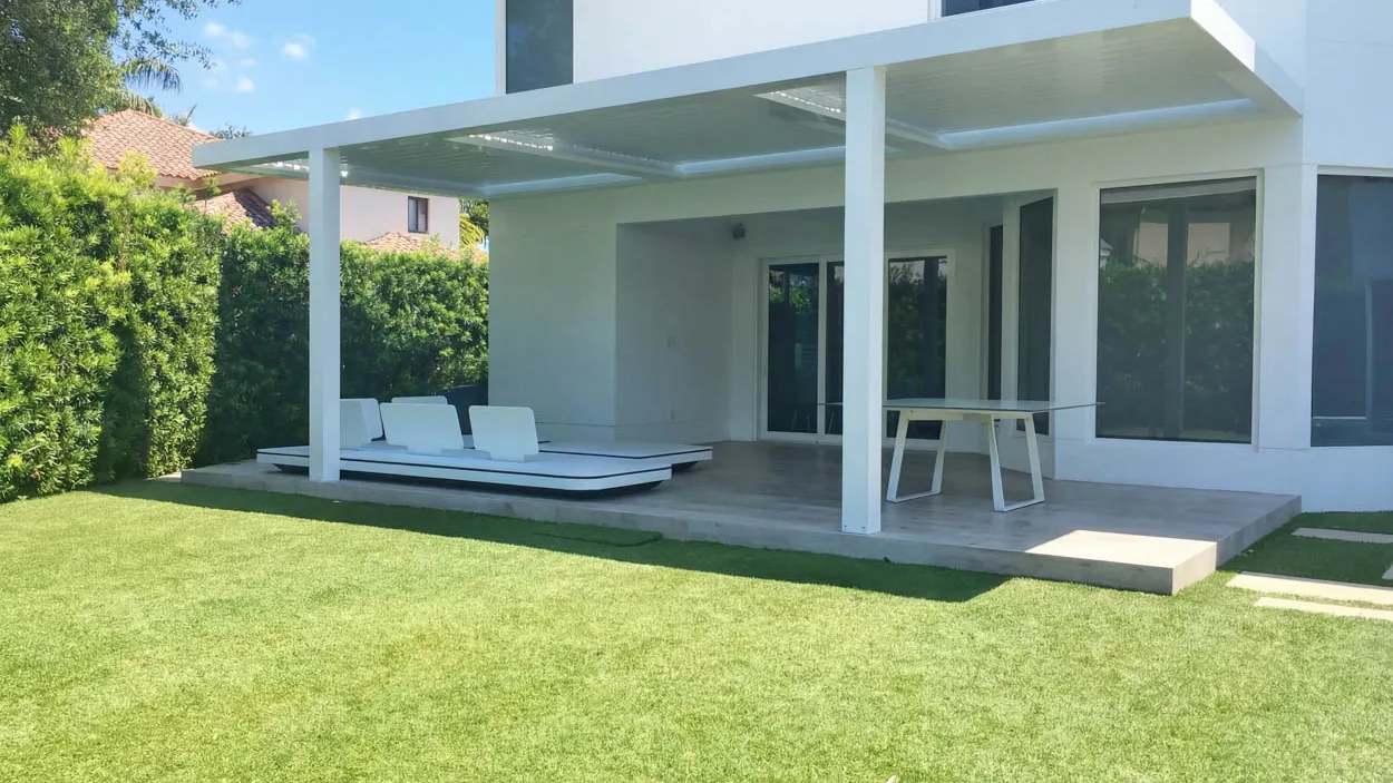 White louvered roof attached to the back of a two-story white house, covering a gray tile terrace with a low white daybed and a dining table, with a tall hedge at left, a neighbor's barrel tile roof beyond it and lawn in the foreground.