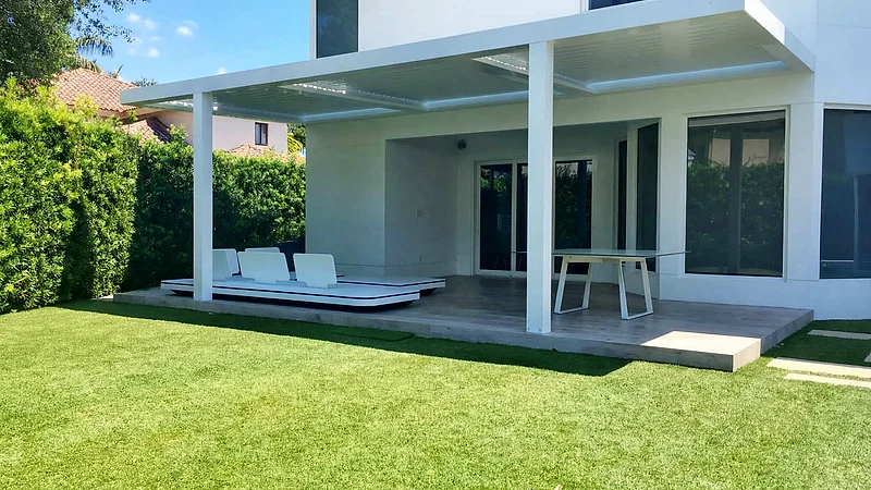 Modern white house with a louvered roof over a raised terrace holding a daybed and a table, beside a tall hedge and lawn