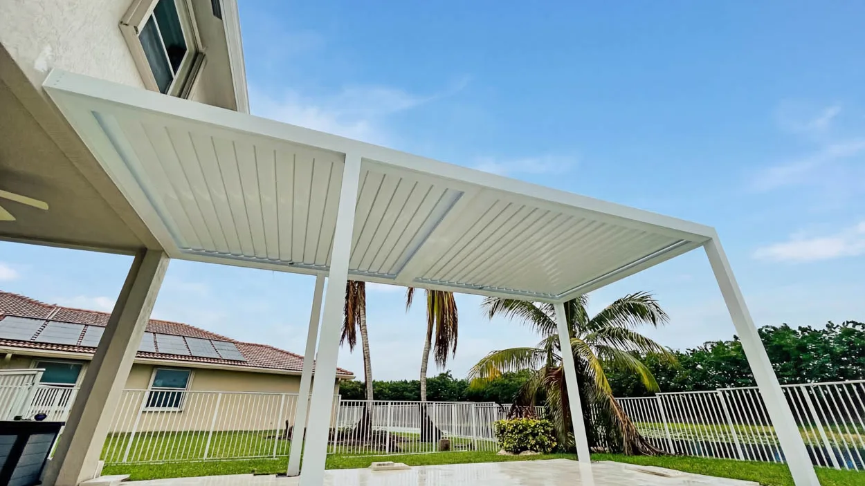 White louvered roof on slim posts over large-format paving still wet after rain, with an aluminum fence, a lake behind it and solar panels on the neighboring tile roof.