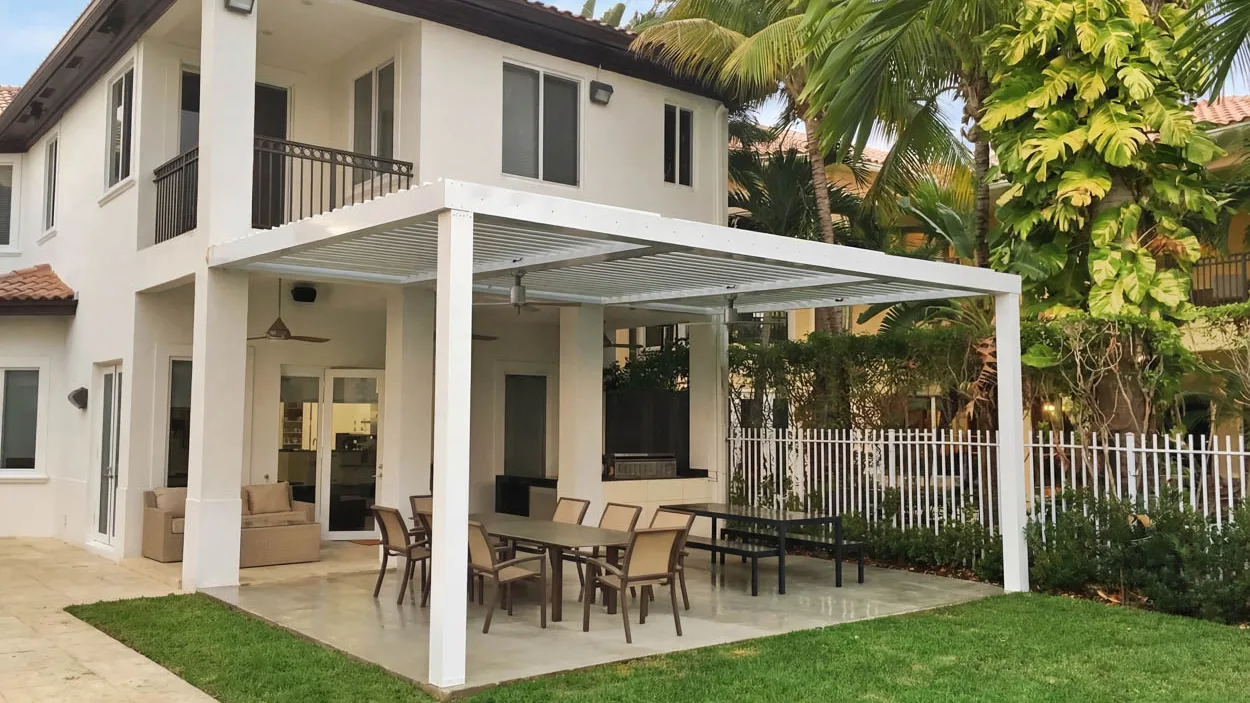 White louvered roof over an outdoor dining table and chairs, built alongside an existing covered porch that already has a ceiling fan mounted under it, with a white picket fence and palms behind.