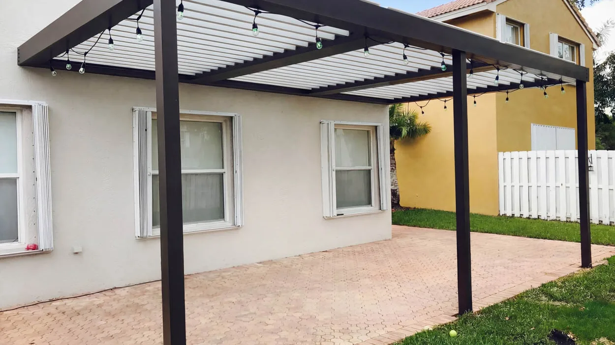 Motorized louvered roof over a paver patio, with aged aluminum shutters on the house windows, a weathered white wood fence behind and a tennis ball left on the lawn.