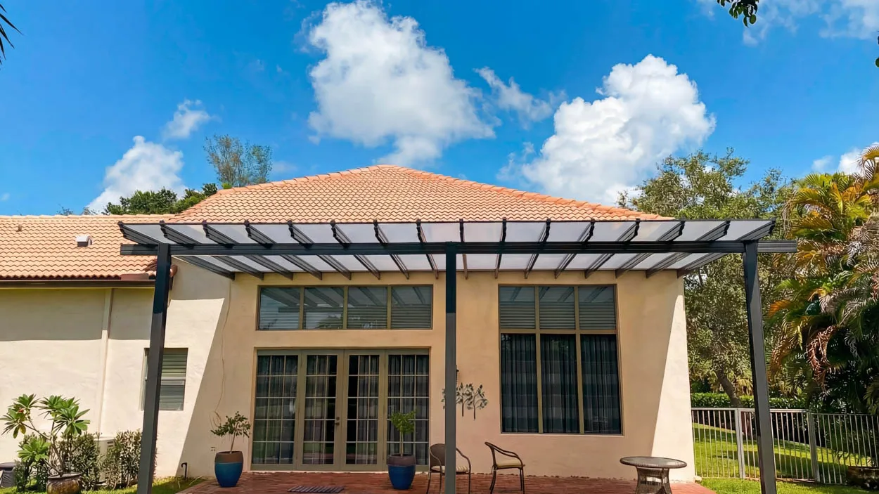 Attached patio cover with translucent roof panels over a red brick paver patio, with two blue planters and a pair of metal chairs in front of the French doors of a tile-roofed stucco house.