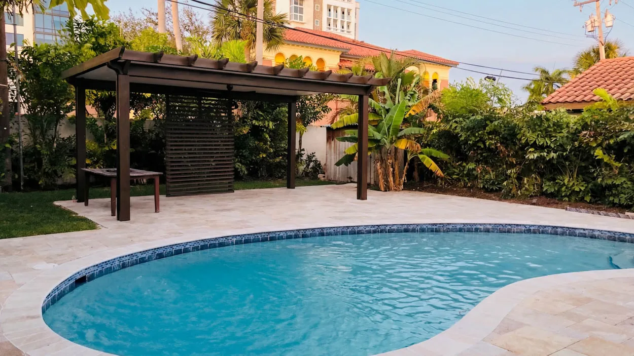 Dark-framed pergola beside a kidney-shaped backyard pool, with overhead power lines crossing above the yard and a wooden louvered privacy screen and a bench underneath it.