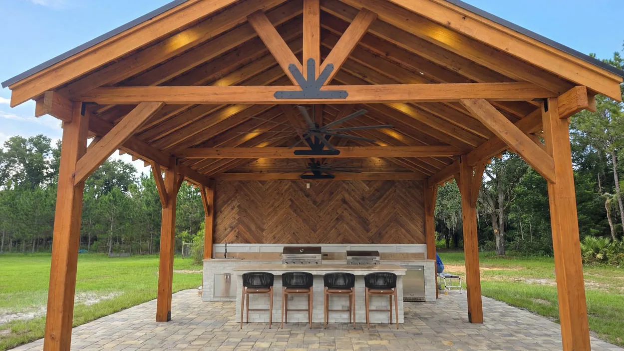 Timber pavilion with a gable roof and exposed trusses bolted with steel plates, covering an outdoor kitchen with two built-in grills, a stone island with bar stools and ceiling fans hung along the ridge.