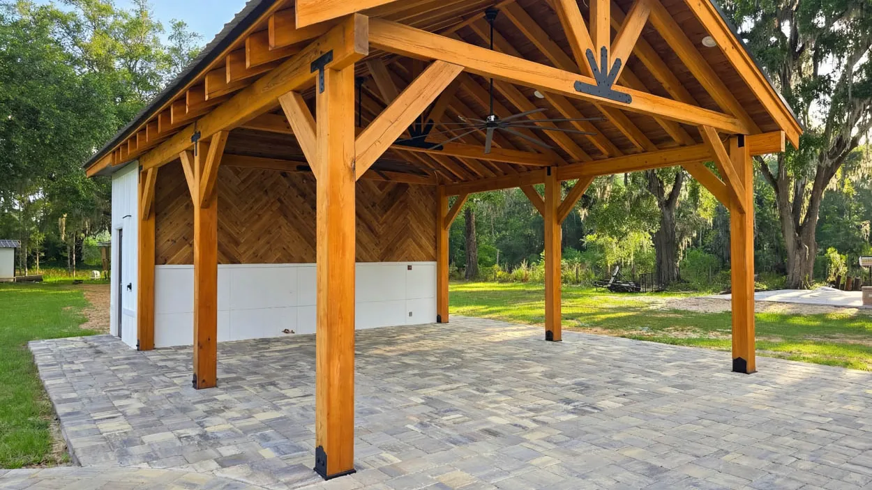 Timber pavilion on a paver patio, its exposed trusses joined by black steel plates and its posts set on black metal bases, built against a white outbuilding with live oaks draped in Spanish moss behind.
