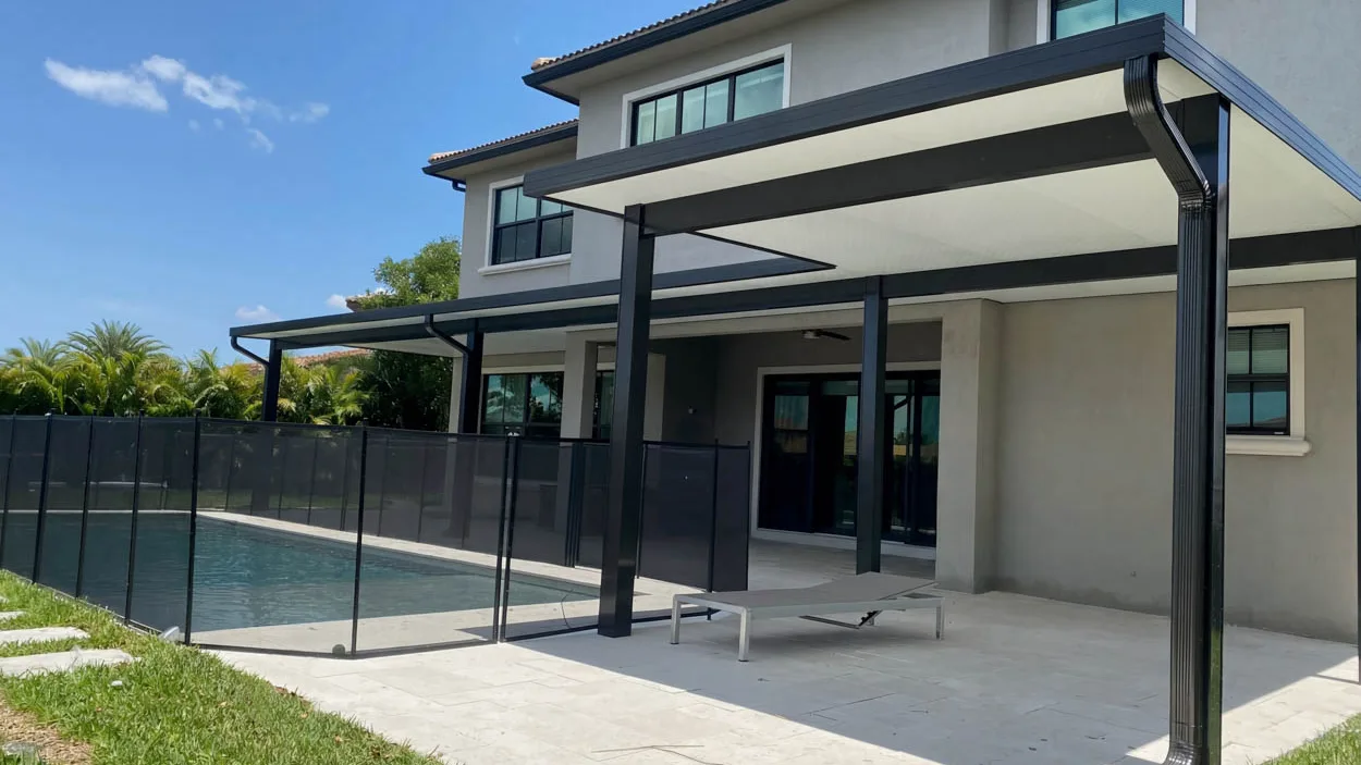 Aluminum patio cover next to a pool, with a removable mesh safety fence, sheets of construction plastic on the grass and a ribbed downspout at the corner of the house.