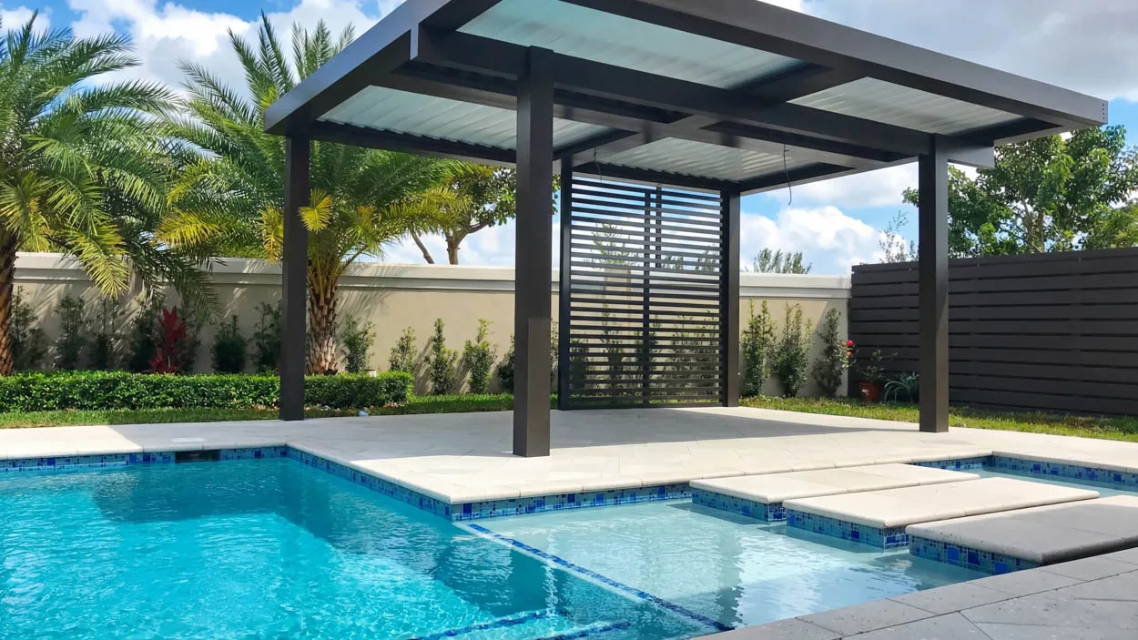 Freestanding dark aluminum pergola with translucent roof panels beside a pool, with a slatted privacy panel under it and two thin black cables left hanging from the frame.