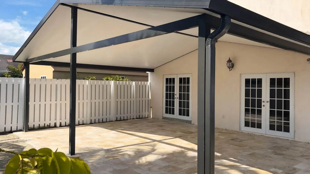 Gabled aluminum patio cover attached to a stucco house, with a weathered wall lantern beside it and a white PVC fence behind.
