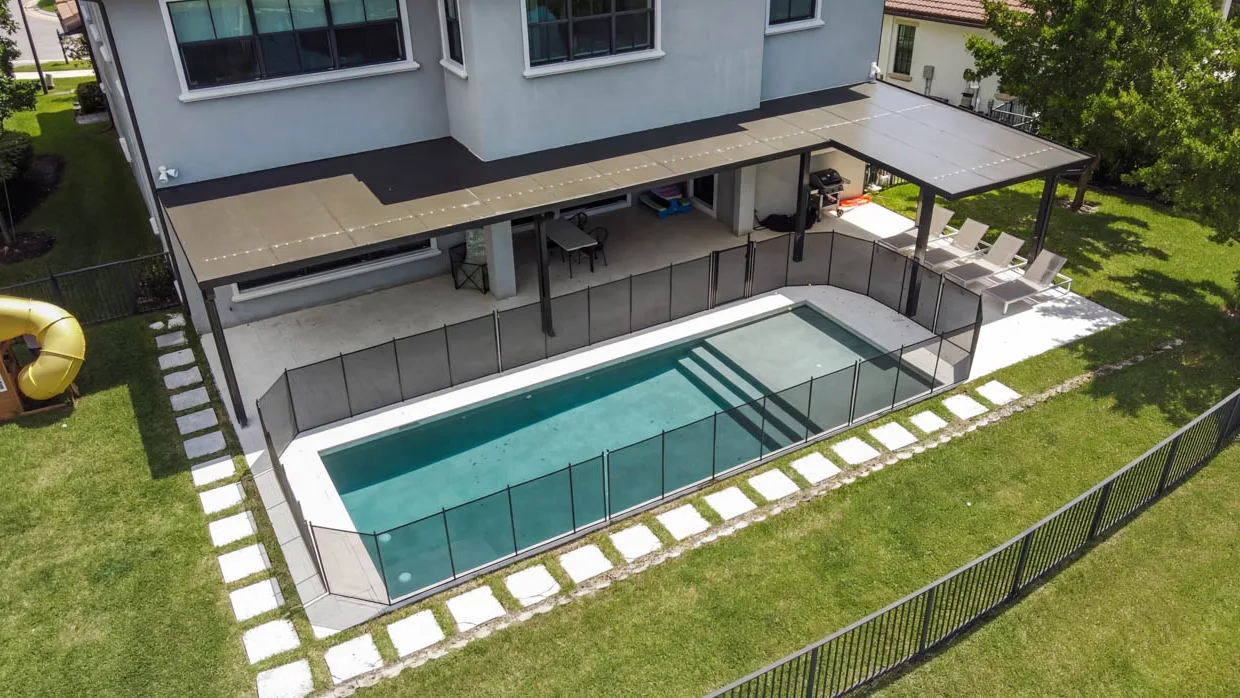 Aerial view of a pool behind a house, ringed by removable mesh safety fencing, with a yellow plastic slide, a gas grill and an orange kayak on the lawn.