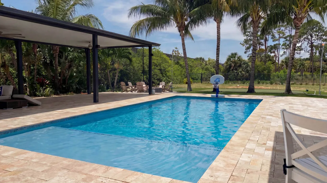 Rectangular pool with a shallow shelf at the near end and a travertine paver deck, a basketball hoop at the far edge, a covered lanai with ceiling fans to the left and palms behind a black metal fence.