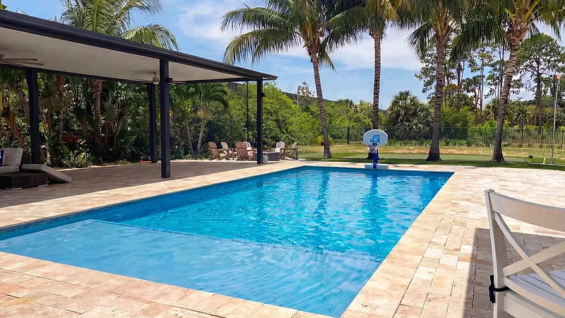 Pool on a travertine deck beside a covered patio on dark posts, with palms, lawn and an open field beyond