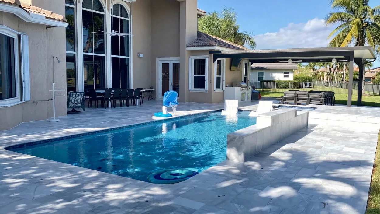 Pool with a blue float in the water beside a marble deck, an outdoor shower on the house wall and a dining table under tree shadows.