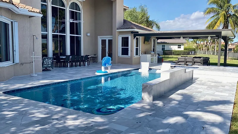 Pool with in-water loungers beside a raised marble-look wall, a covered lounge area and a two-story house with arched windows