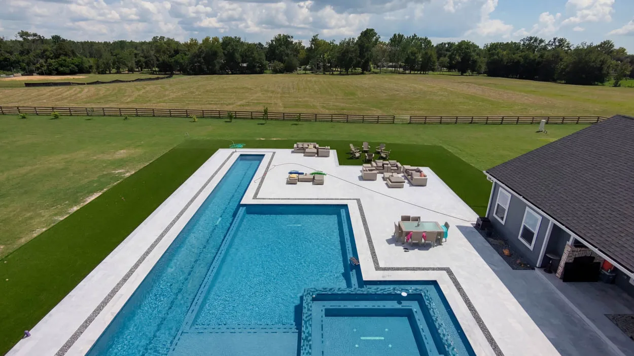 Aerial view of a long pool and a large square spa on a wide deck, with a garden hose and its reel crossing the deck, groups of lawn furniture and a wood ranch fence around the pasture behind.