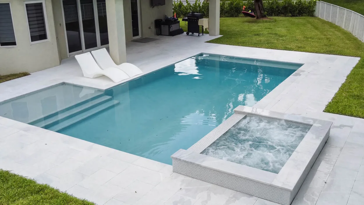 Raised spa clad in small white tile running with its jets on at the corner of a rectangular pool, with two white in-water loungers on the shallow ledge and a grill under the covered lanai.