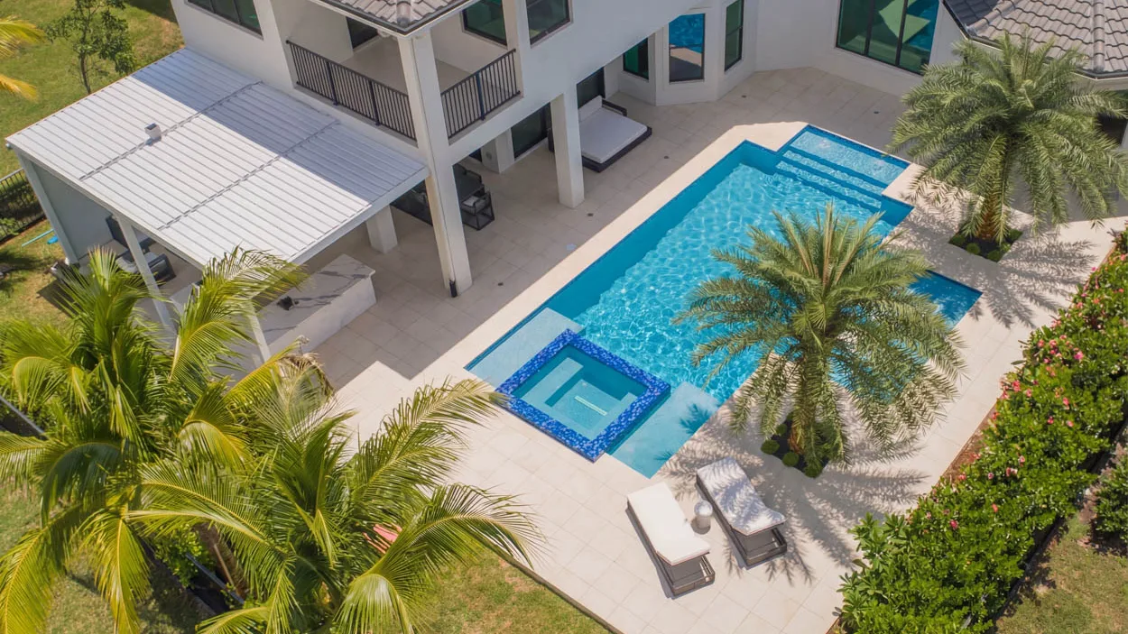 Aerial view of a backyard pool with a robotic cleaner on the floor, a daybed under the porch and an outdoor kitchen beneath a metal roof.