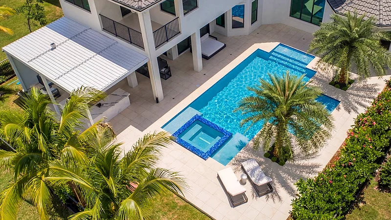 Aerial view of a pool and square spa on a pale stone deck beside a white louvered roof, a two-story house and palms