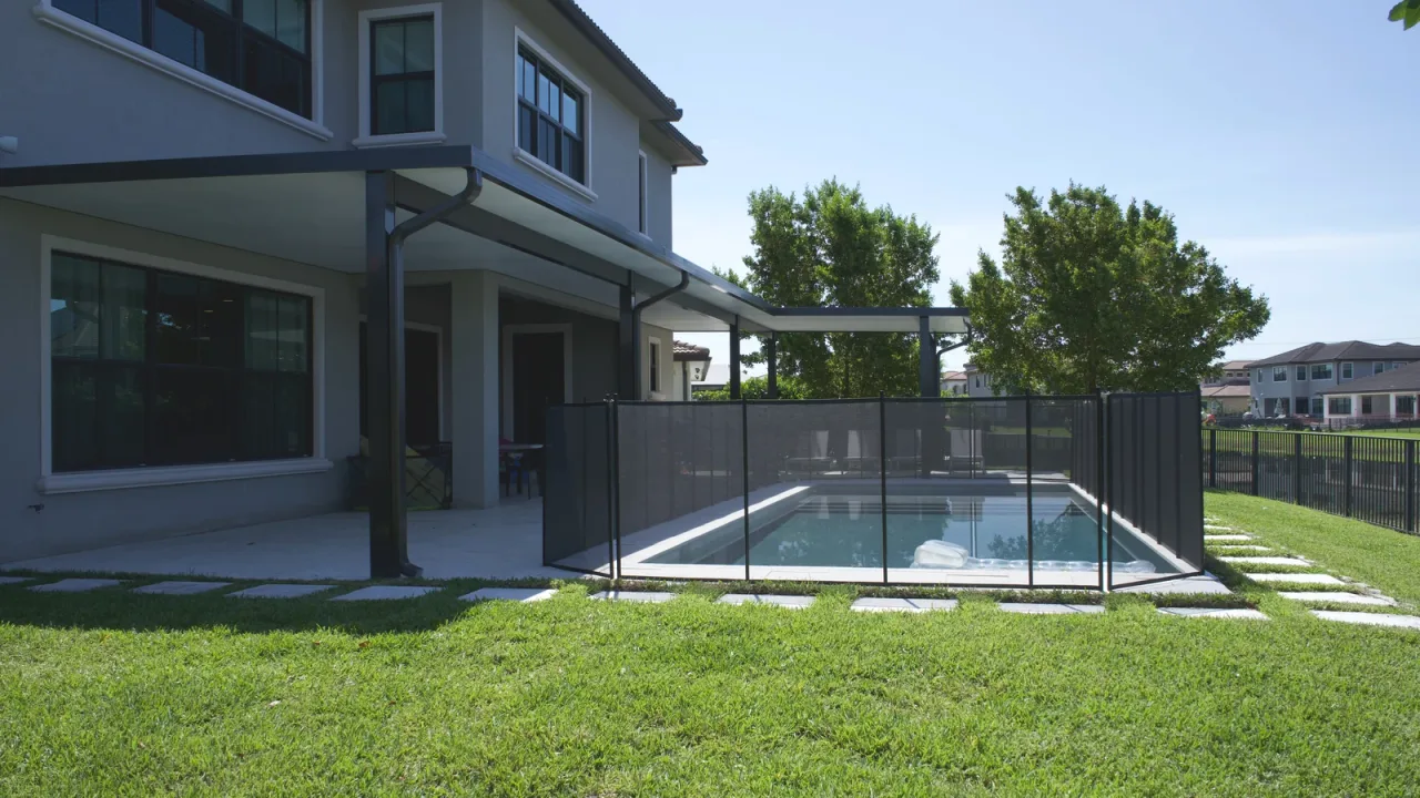 Rectangular pool behind a two-story house, enclosed by a black mesh safety fence, with a dark-framed patio cover alongside.