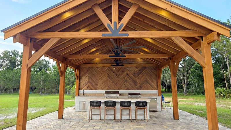 Timber pavilion with a vaulted roof and ceiling fan, sheltering an outdoor kitchen and bar seating.
