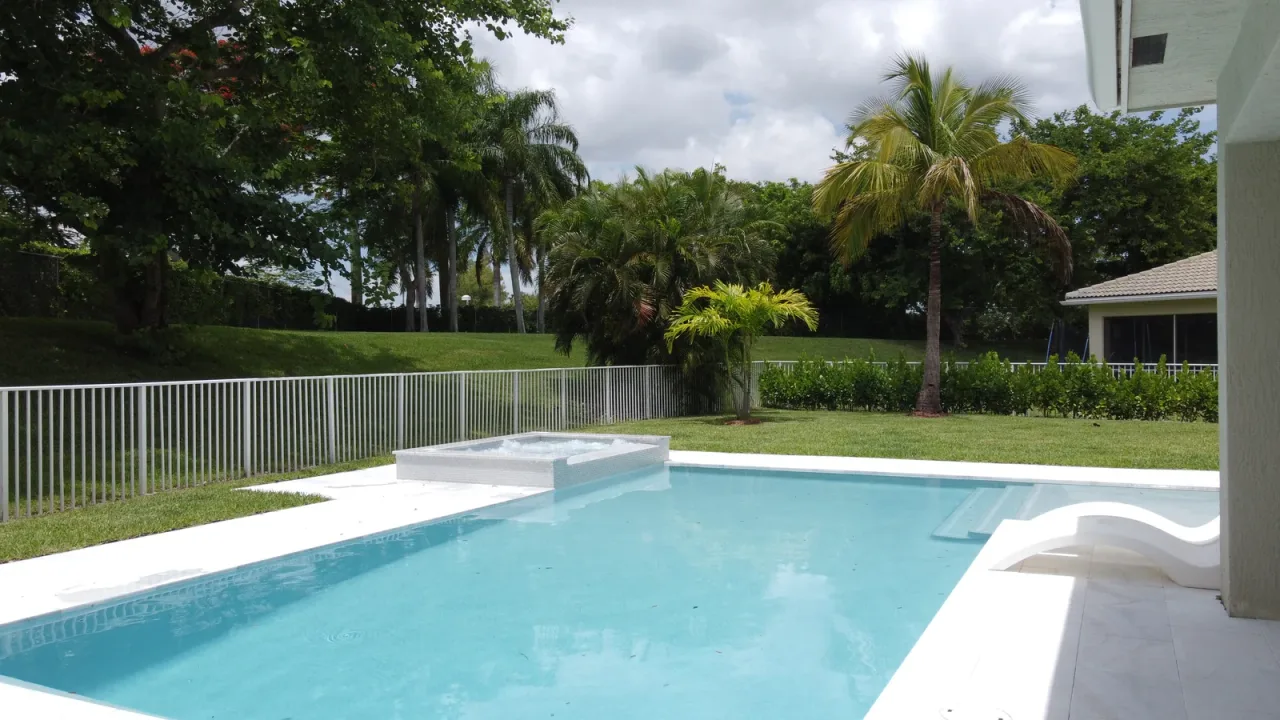 Pool with a raised spa bubbling at its far edge, white coping, a white lounger by the steps and trees behind a white fence.