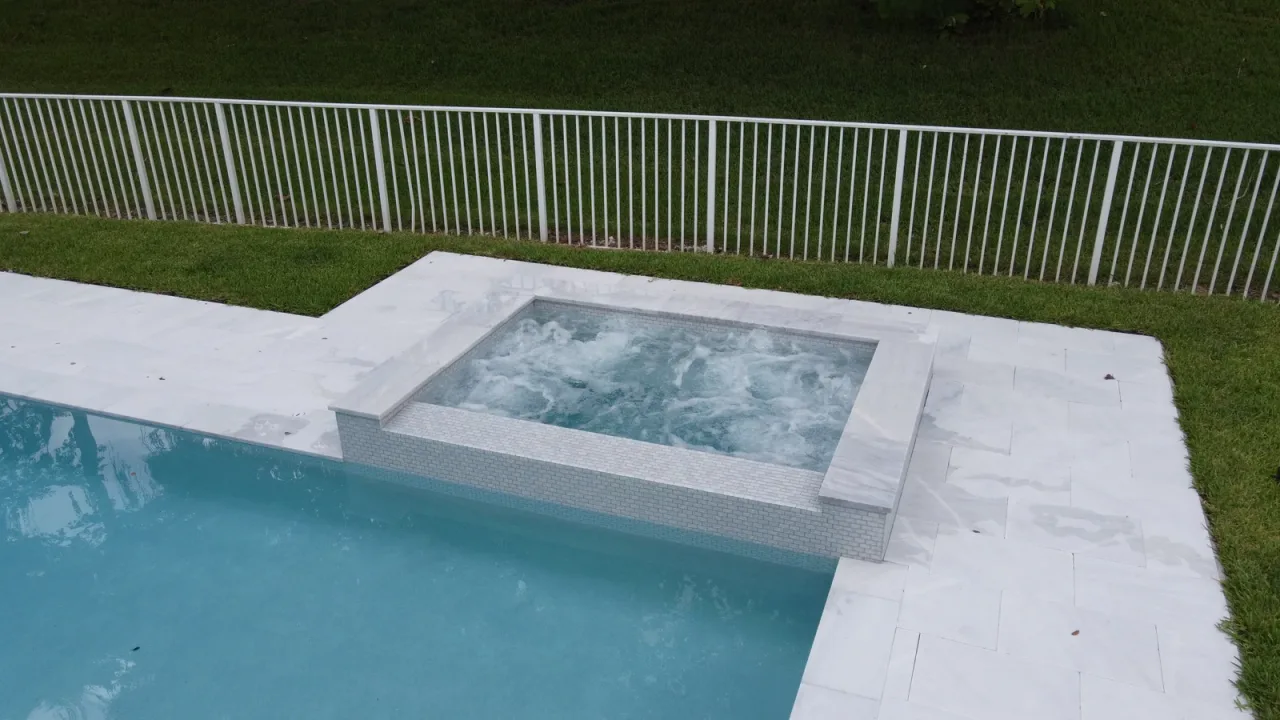 Square spa with its jets running, raised slightly above the pool and edged in white stone, with a white metal fence behind.
