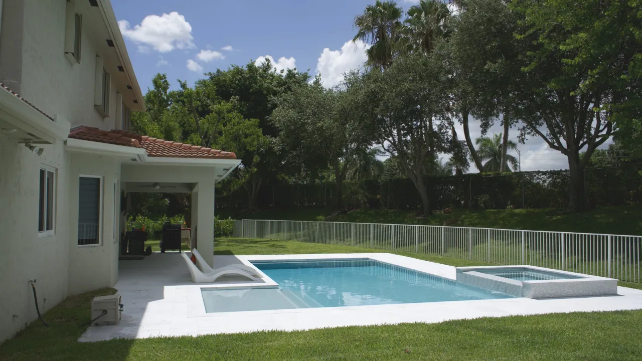 Light blue pool and raised spa beside a covered lanai, with two white loungers at the corner and trees behind a white fence.