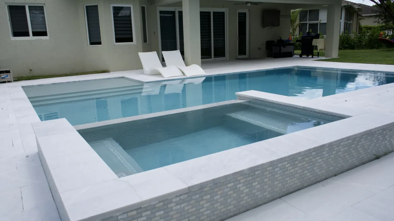 Raised spa clad in pale mosaic tile, with an inside bench, beside a pool and two white loungers near a covered lanai.