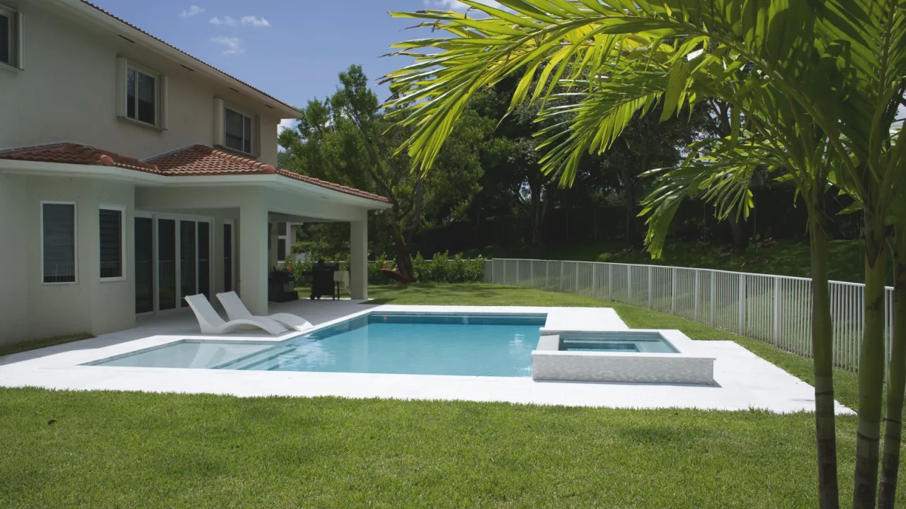Backyard pool with a raised spa and white stone deck beside a two-storey house, with loungers, lawn and a palm.