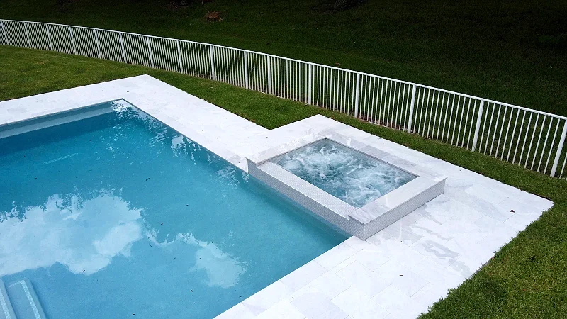 Aerial view of a rectangular pool with a raised mosaic-clad spa and a wide white stone deck in a fenced Florida backyard.