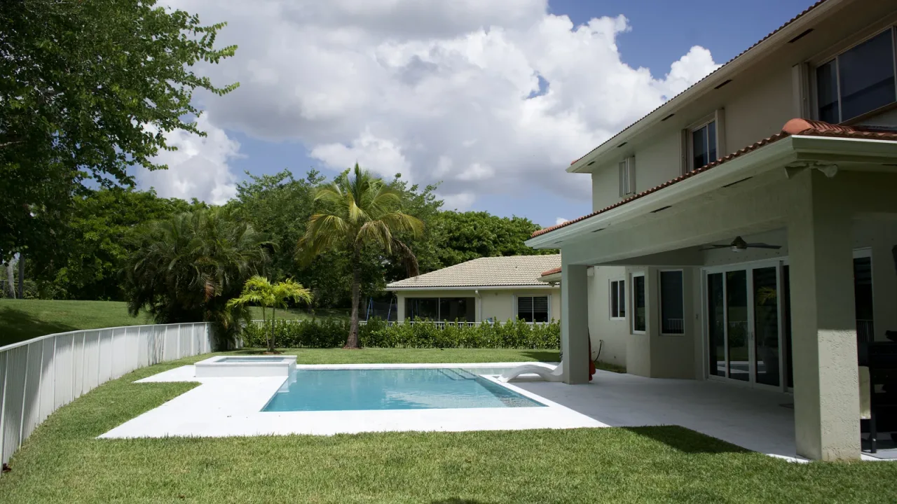 Rectangular pool with a raised spa and white deck beside a two-storey house, framed by lawn, palms and a white fence.