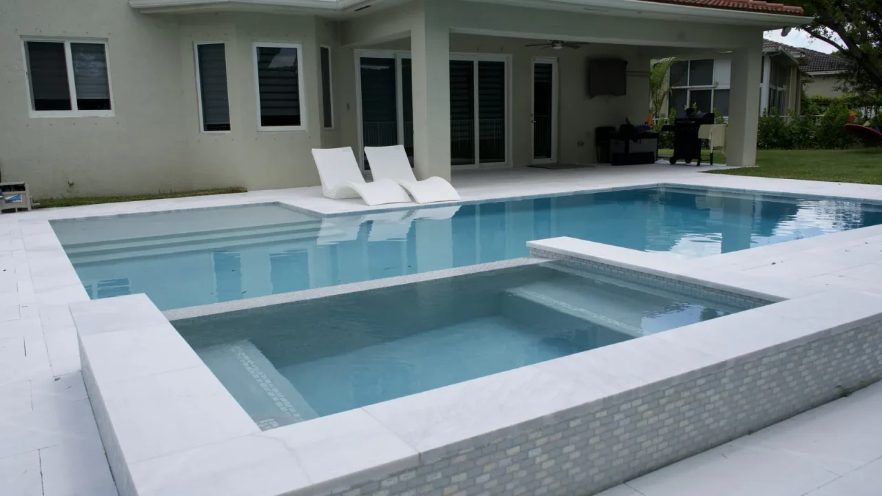 Raised spa wrapped in pale mosaic tile in a light-finish pool, with white stone coping and two loungers on the deck beyond.