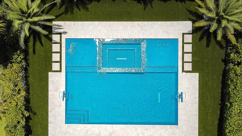 Overhead view of a rectangular pool with an inset spa, bordered by a stone deck, clipped hedge and palms.
