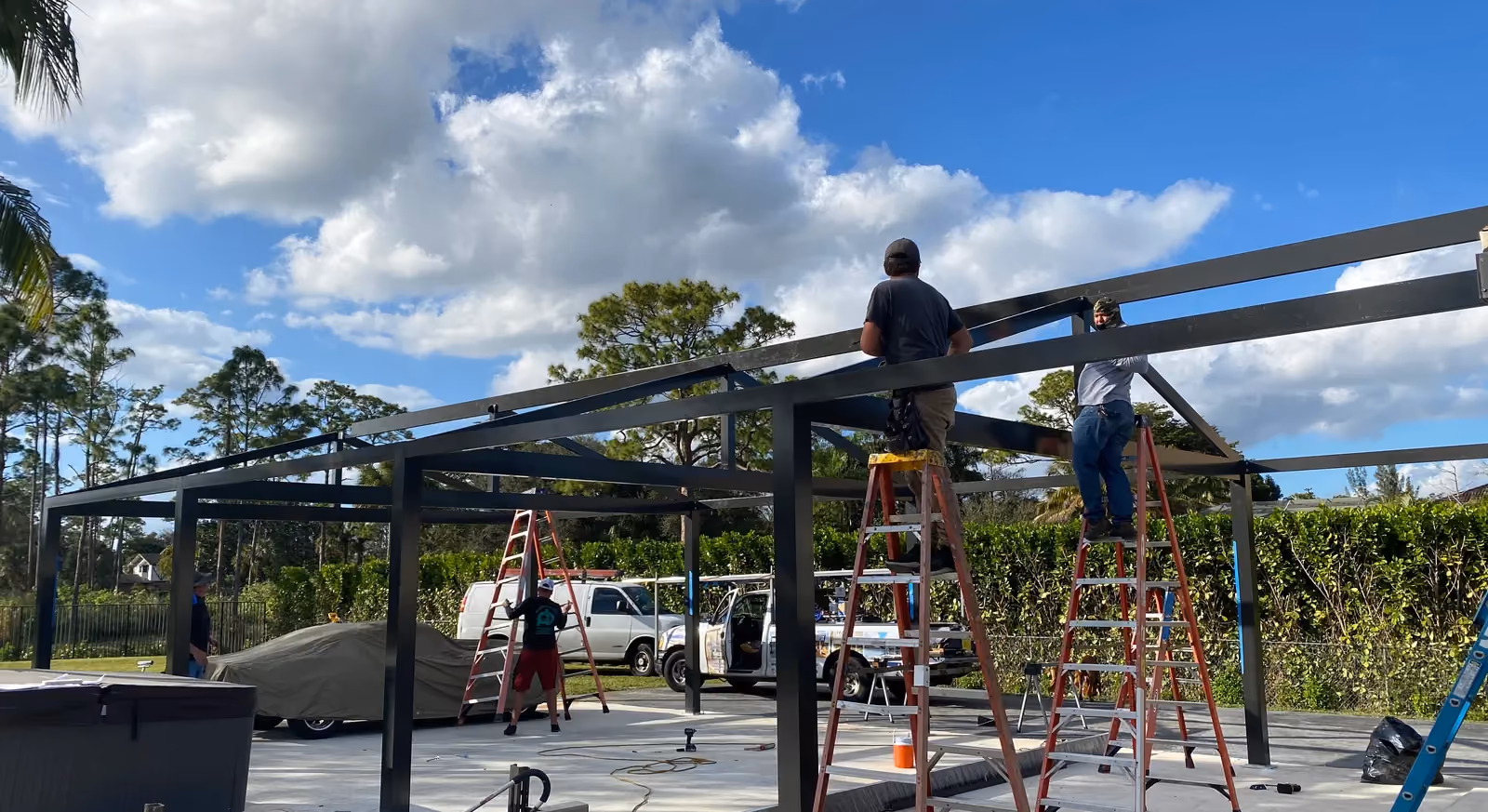 Crew on ladders assembling a black aluminum pergola frame over a travertine patio.