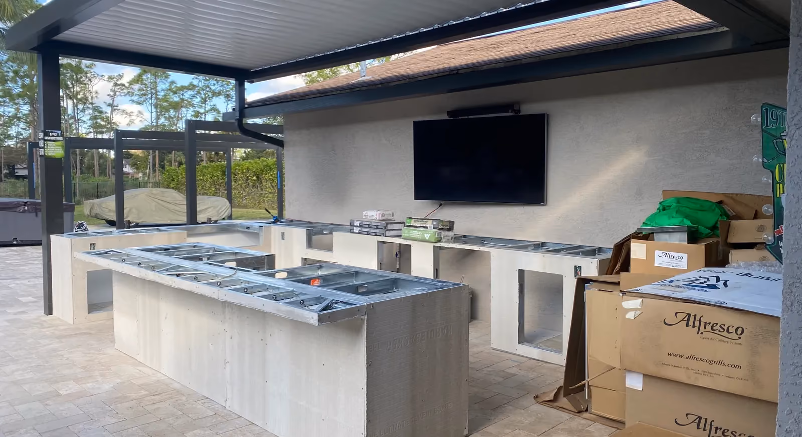 Outdoor kitchen island and counters framed in steel and cement board under a louvered pergola.