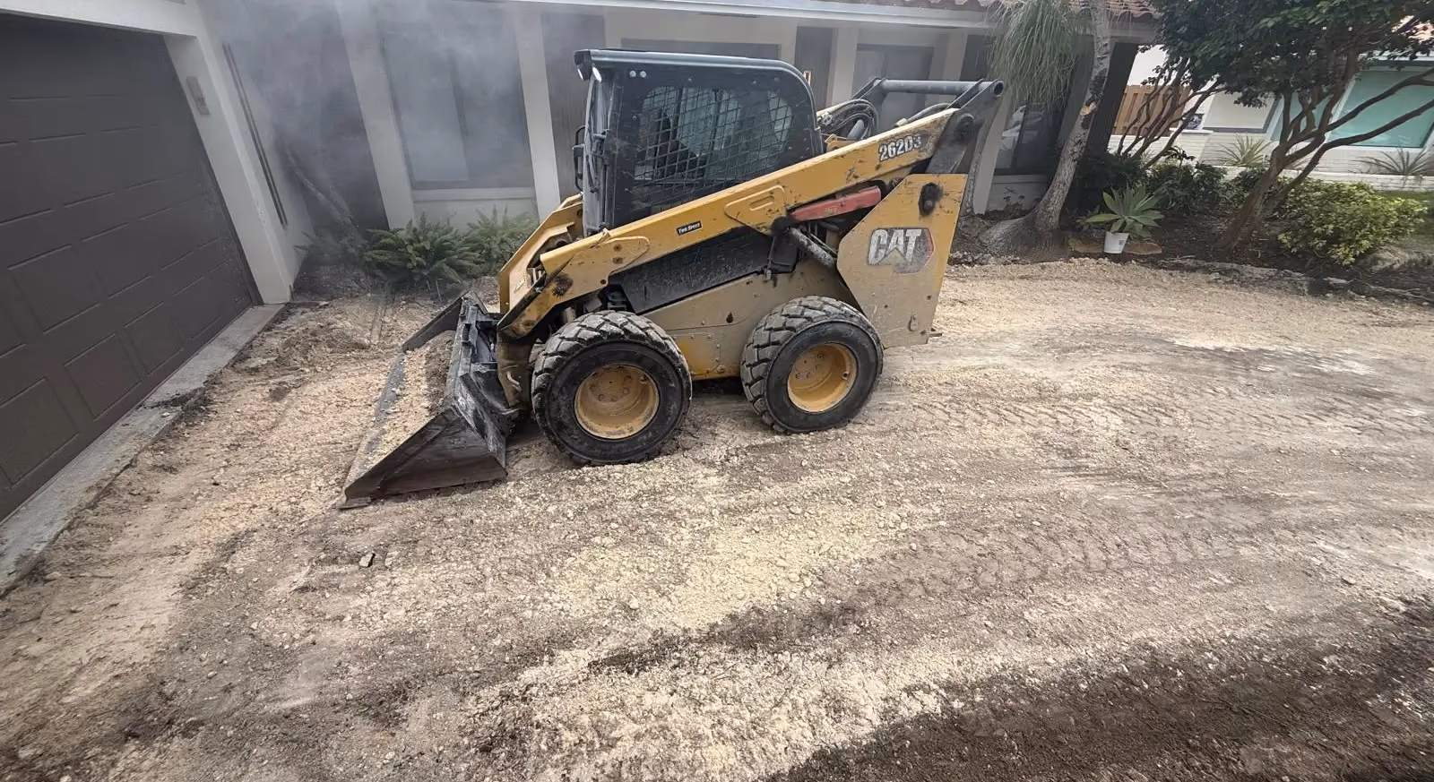 Skid steer grading the base of a demolished driveway in front of a tile-roof house.