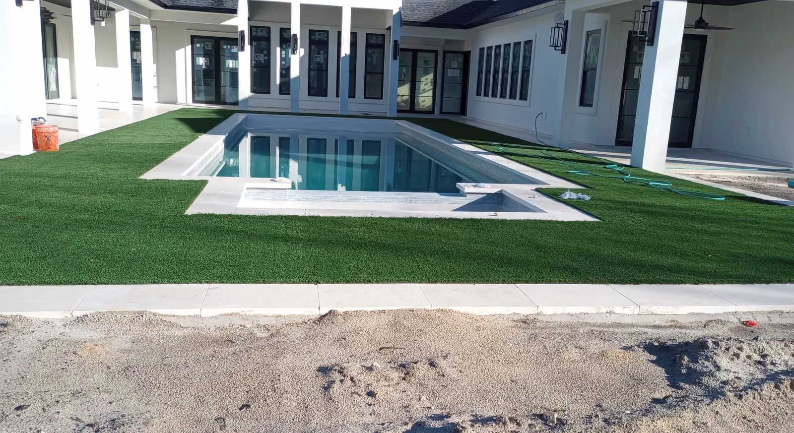 Artificial turf laid around a new rectangular pool with spa in the courtyard of a white home.