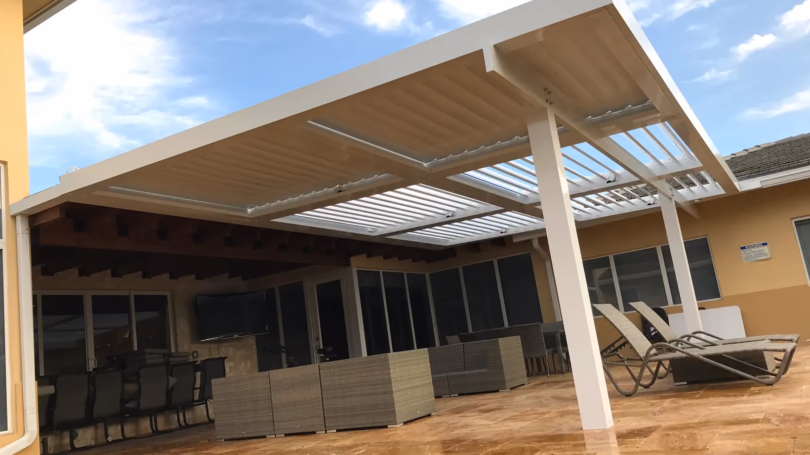 Large white louvered roof with partly open blades over a travertine patio with wicker lounge furniture