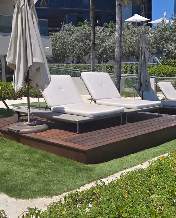 Dark hardwood lounge deck with daybeds set on artificial turf beside dune planting.