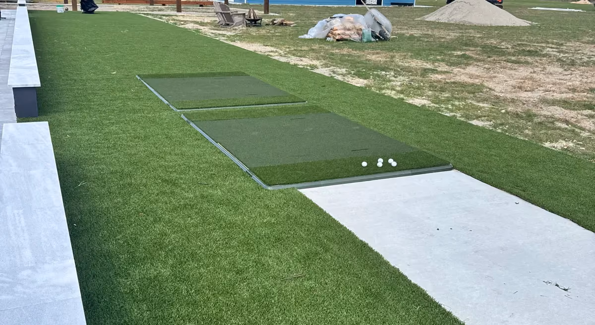 Golf hitting mats set into artificial turf beside a concrete pad, with a blue metal barn in the background.