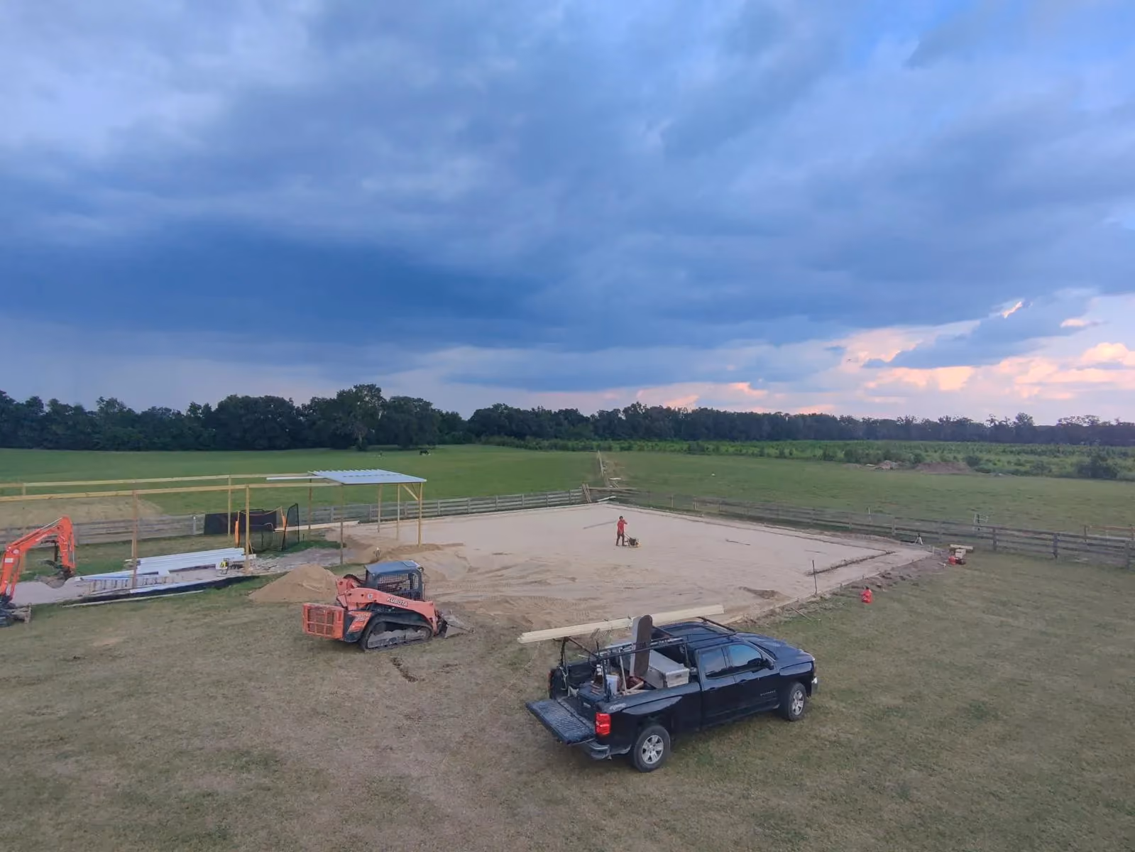 Aerial view of a compacted sand pad being prepared for a concrete slab beside pole framing on a rural lot.