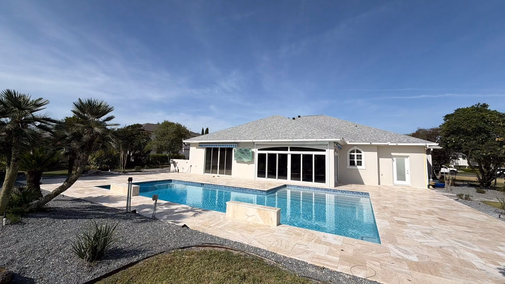 Pool remodeling in Florida: blue glass waterline tile and travertine deck behind a stucco home, by Mr & Mrs Outdoor Living