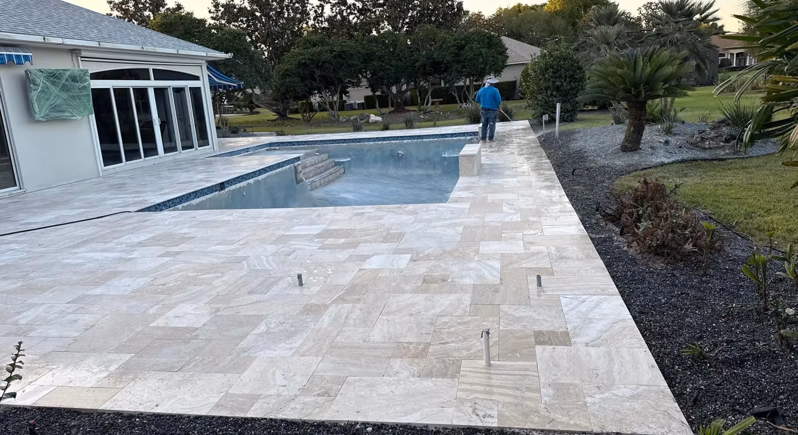 Large French-pattern travertine deck bordered by black lava rock around a pool in progress.