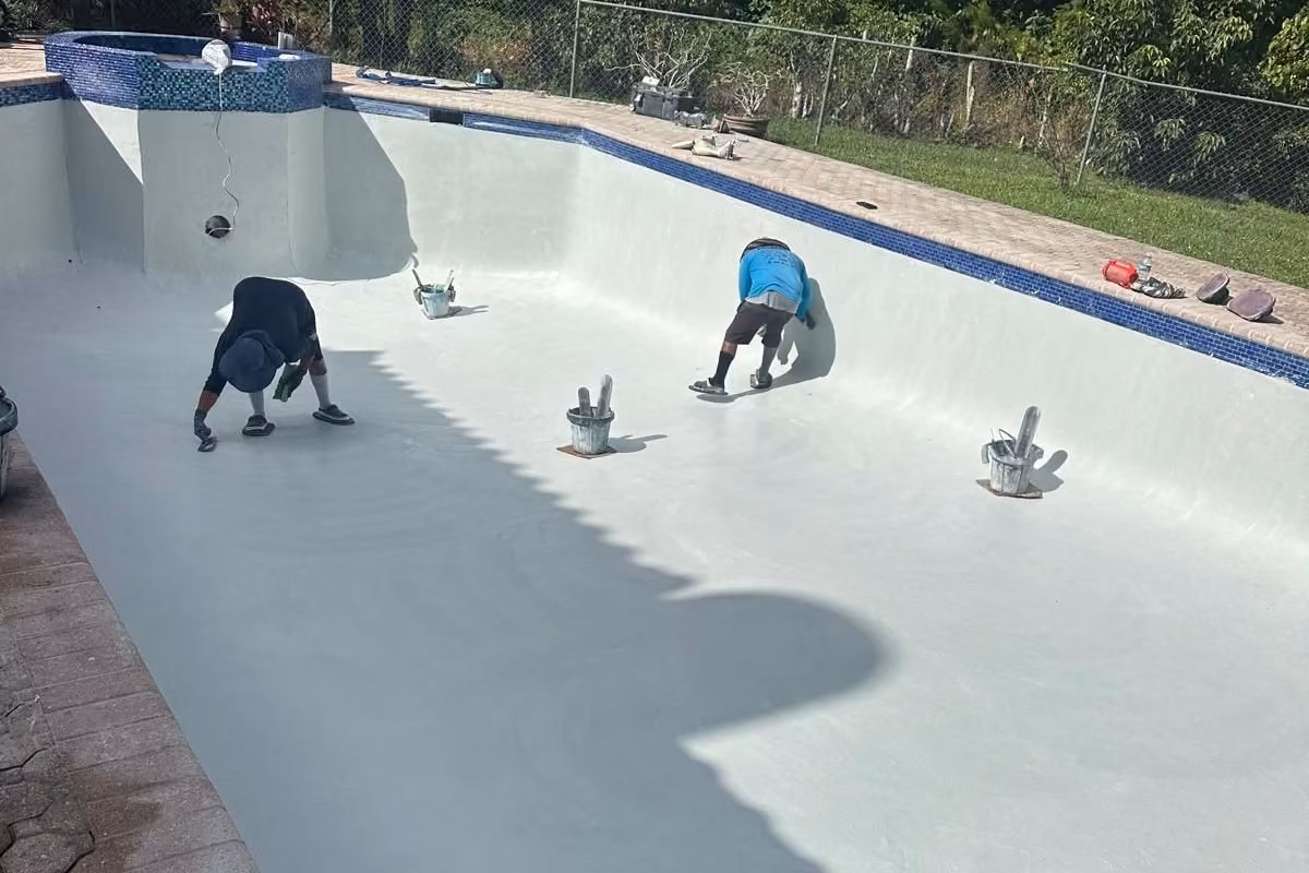 Two workers hand-troweling new white plaster in a drained pool with blue waterline tile, pool renovation in Florida