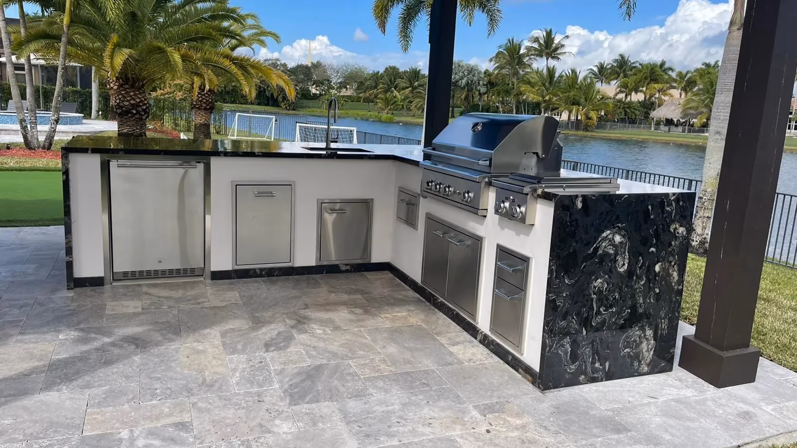 L-shaped outdoor kitchen with black granite waterfall under a dark pergola beside a lake.