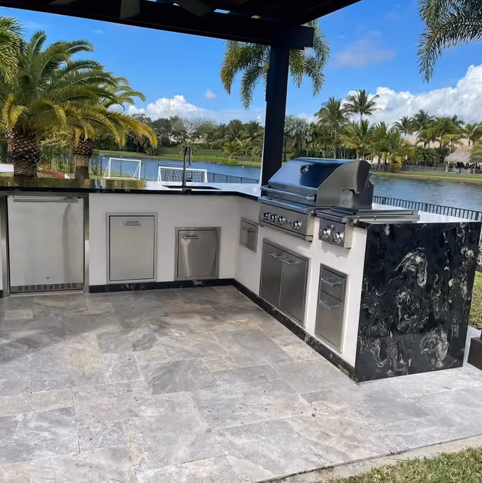 L-shaped outdoor kitchen with black granite waterfall under a dark pergola beside a lake.