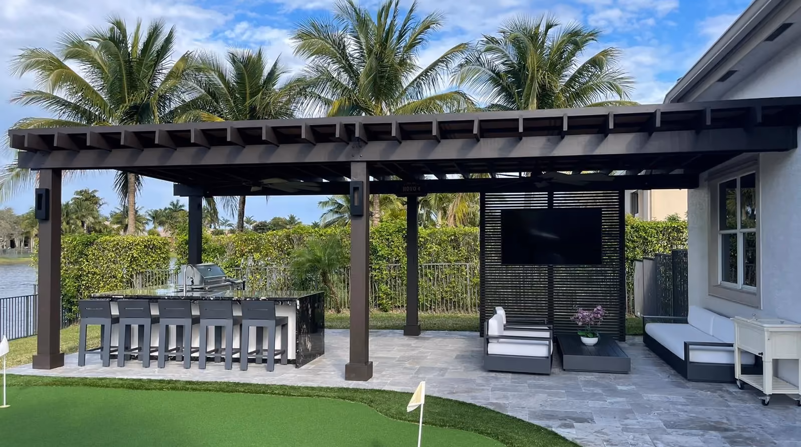 Dark wood pergola over an outdoor kitchen bar and lounge with TV screen beside a putting green.